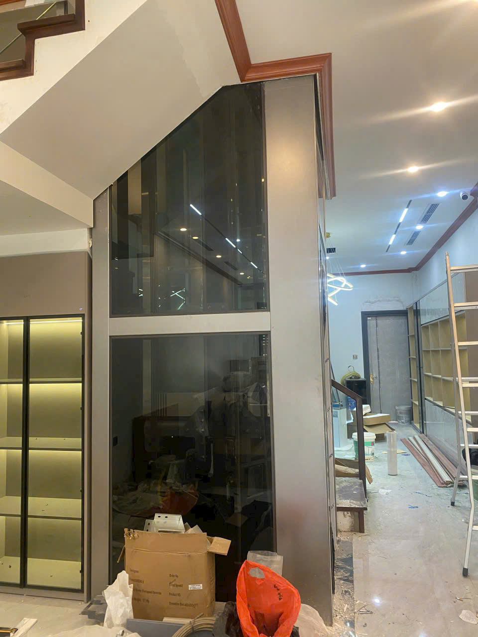 Thang máy kính gia đình đang lắp đặt trong nhà, xung quanh còn ngổn ngang vật liệu thi công